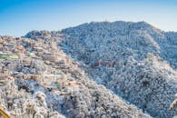 A scenic view of Shimla in winter, with snow-covered buildings and forests under a clear blue sky.